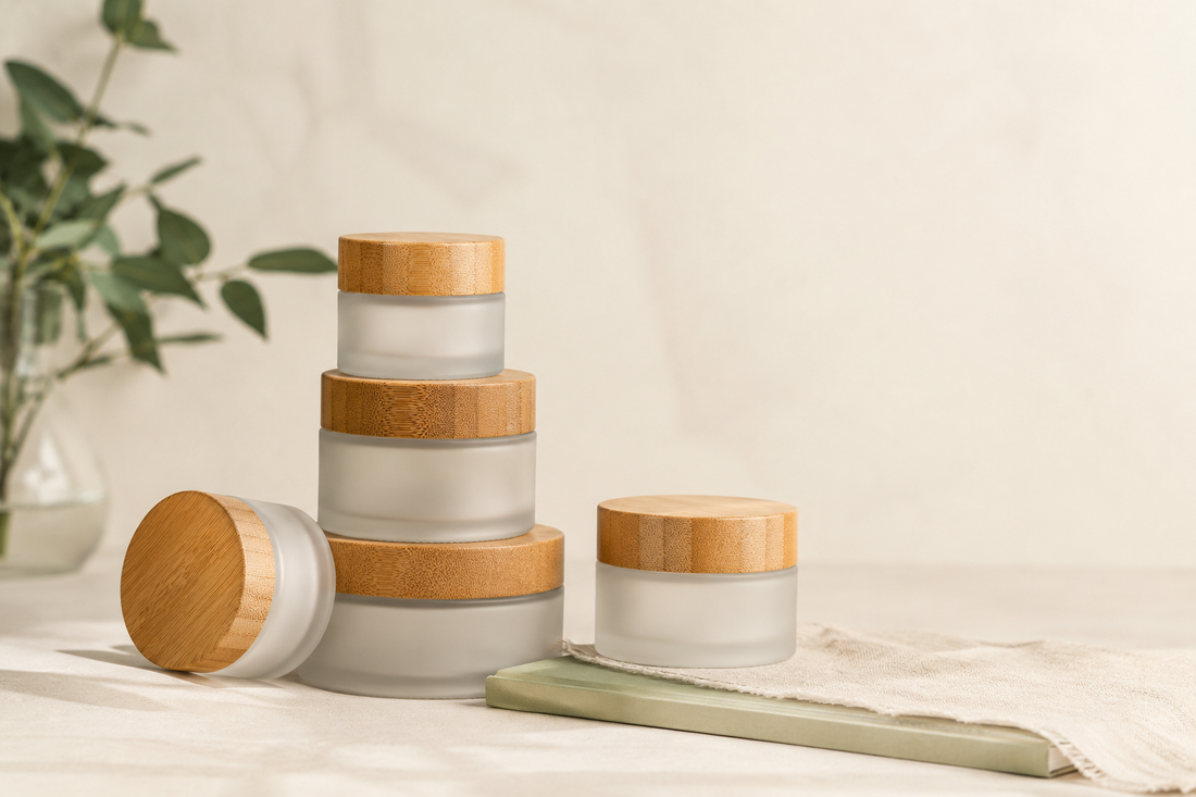 Frosted glass skincare jars with wood lids stacked on a cream surface beside linen, sage accents, and soft greenery.