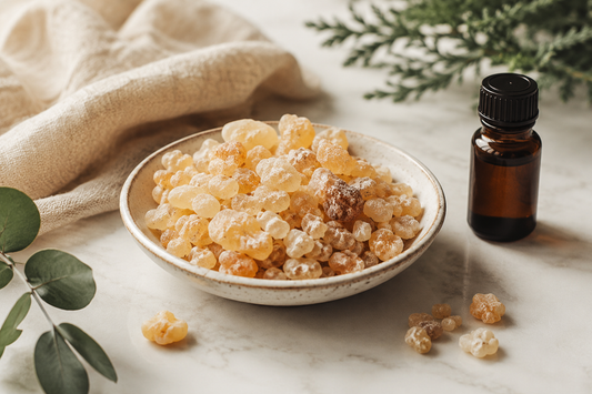 Bowl of golden frankincense resin beside an amber essential oil bottle on a cream stone surface with linen and greenery.
