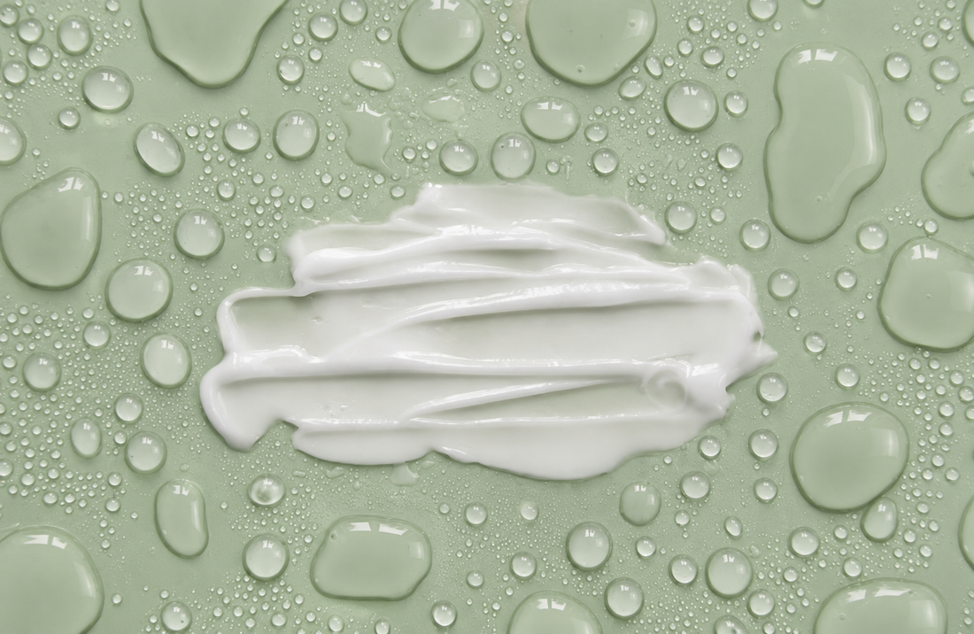 White cream smear on a sage green surface covered with glossy water droplets.