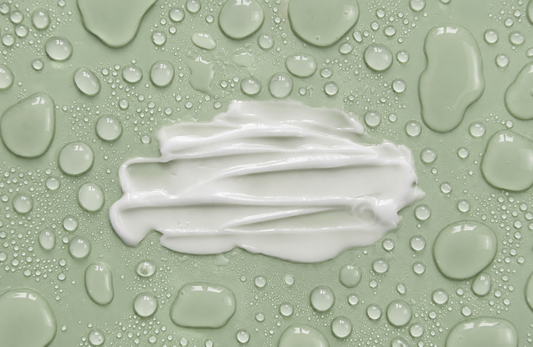 White cream smear on a sage green surface covered with glossy water droplets.