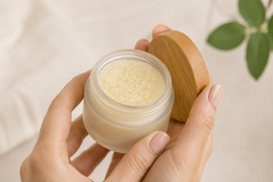 Feminine hands holding an open frosted jar of grainy tallow balm with a wood lid against a soft cream background.